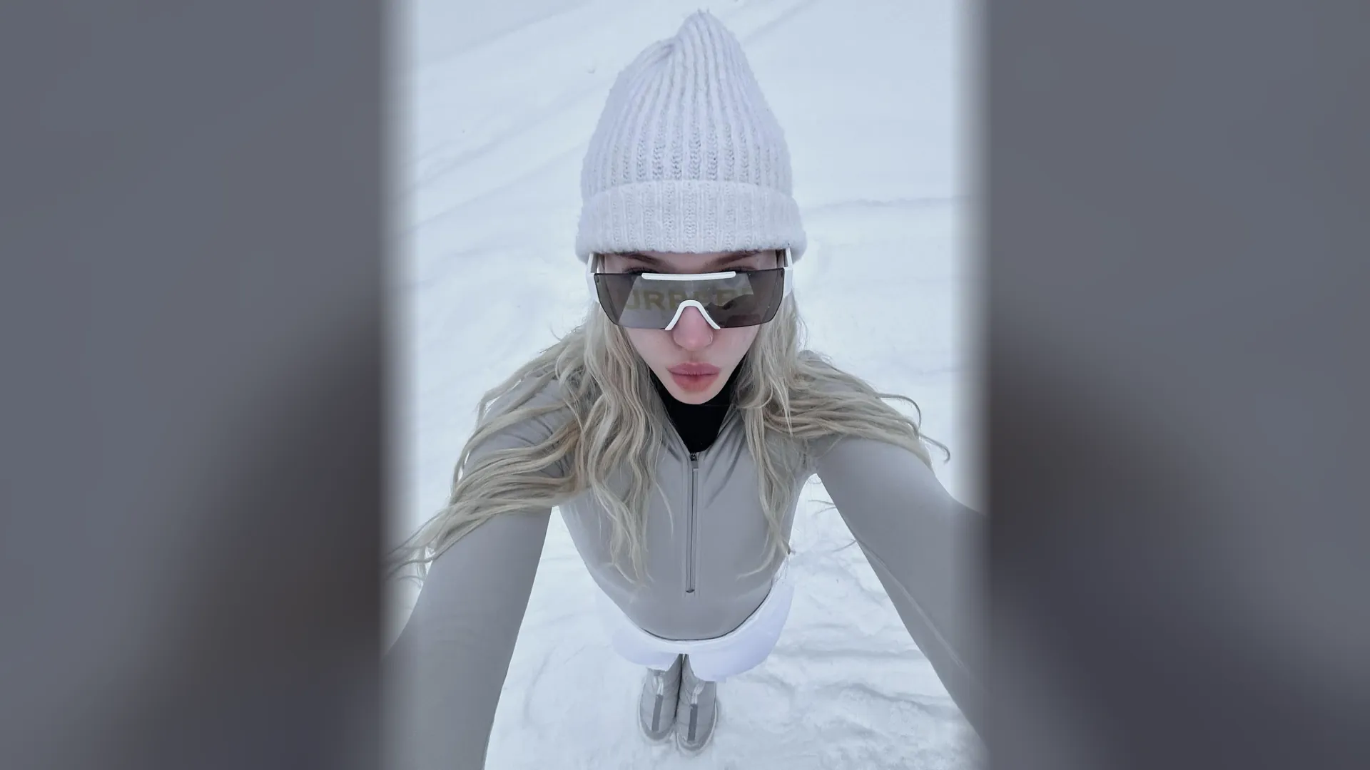 Alexandra Marianna taking a wide-angle selfie in the snow, in a white beanie and shield sunglasses