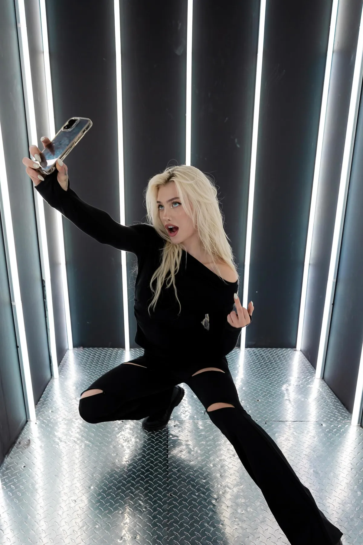 Alexandra Marianna dressed in black in a corridor of light tubes, holding her phone