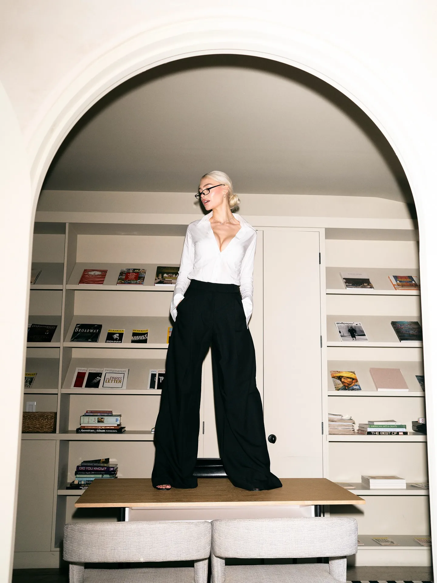 Alexandra Marianna in a white shirt and black trousers, standing in an arched doorway lined with bookshelves